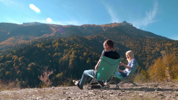 Chico y chica están sentados en asientos plegables bebiendo té y mirando las montañas — Vídeo de stock
