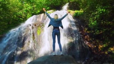 Genç, narin sarışın bir kayanın üzerinde duruyor ve tropik bir şelale düşünüyor.