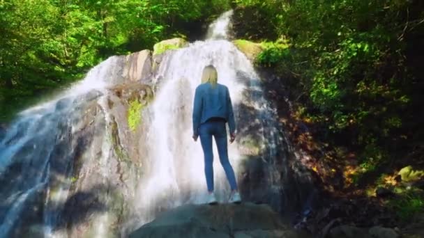 Giovane bionda snella in piedi su una roccia, contempla una cascata tropicale — Video Stock