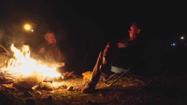 Young guy and a girl are sitting on folding chairs by the fire and talking. — Stock Video