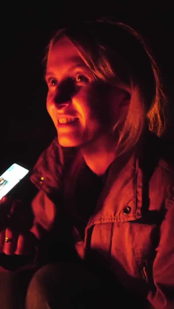 La fille est assise près du feu et envoie des SMS sur les réseaux sociaux avec son téléphone. — Video