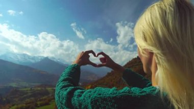 Dağın tepesinde yürüyüş yapan kadın elleriyle kalp şeklinde parmak çerçevesi yapıyor..