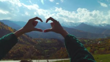 Dağın tepesinde yürüyüş yapan kadın elleriyle kalp şeklinde parmak çerçevesi yapıyor..