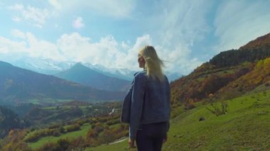 Kot ceketli kadın dağın tepesinde duruyor ve ellerini havaya kaldırıyor.,