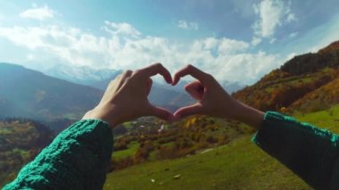 Dağın tepesinde yürüyüş yapan kadın elleriyle kalp şeklinde parmak çerçevesi yapıyor..
