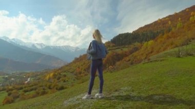 Kot ceketli kadın dağın tepesinde duruyor ve ellerini havaya kaldırıyor.,