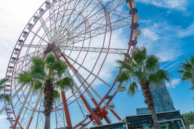 Dönme dolaba yakın çekim, parlak mavi gökyüzüne karşı palmiye ağaçları güneşli bir günde bulutlar, Batumi