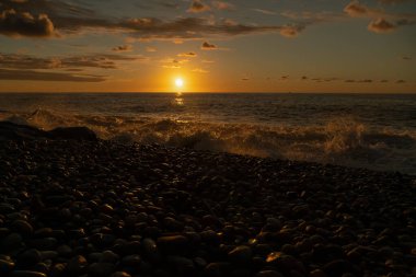 Gün batımında deniz dalgaları çakıl taşlı sahilde yuvarlanır. Güneşin ufukta battığı güzel turuncu bir gün batımı, bir çakıl taşı plajı. Güzel günbatımı arka planı, deniz manzarası