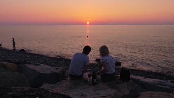 Birbirine aşık bir çift, deniz manzaralı taş bir iskelede oturmuş kırmızı şarap içiyor.. — Stok video