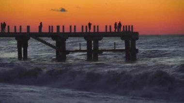 İskelenin arka planında gün batımı İnsanların siluetleri yürür.