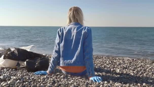 Una mujer en un traje de mezclilla está sentada en la orilla del mar después de la recolección de basura, — Vídeo de stock
