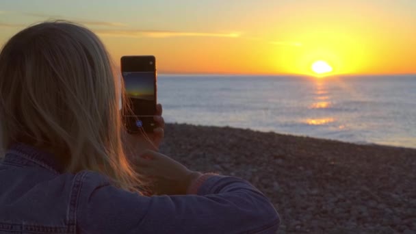 Las manos de las mujeres sostienen un teléfono inteligente y toman fotos de la puesta de sol en el mar. — Vídeo de stock