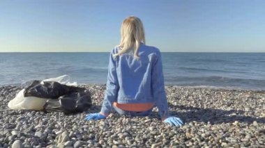 Kot takım elbiseli bir kadın, çöpleri topladıktan sonra deniz kıyısında oturuyor.,
