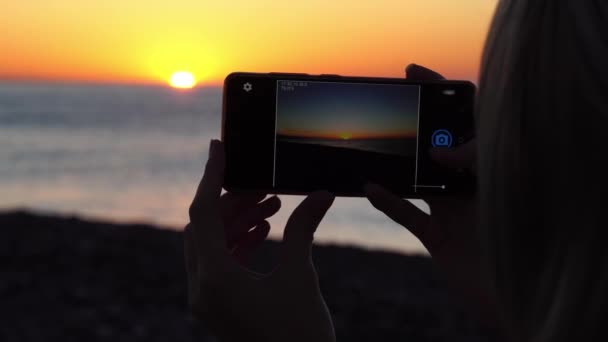 Τα χέρια των γυναικών κρατούν ένα smartphone και φωτογραφίζουν το ηλιοβασίλεμα στη θάλασσα. — Αρχείο Βίντεο
