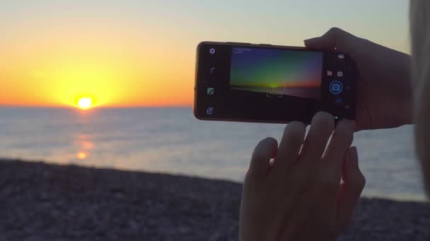 女人的手拿着智能手机，对着海面上的落日拍照. — 图库视频影像