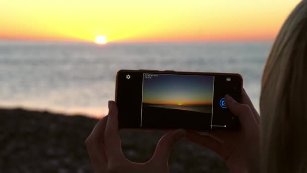 Las manos de las mujeres sostienen un teléfono inteligente y toman fotos de la puesta de sol en el mar. — Vídeo de stock