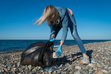 Kadın, güneşli bir günde denizin arka planında siyah bir çöp torbasında plastik, köpük, kauçuk topluyor. Kıyı bölgesinin temizlenmesi. Ekoloji kavramı, gönüllülük.