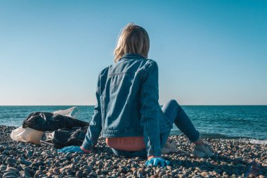 Dünya Çevre Günü. Kadın gönüllü, güneşli bir günde içi çöp dolu çantalarla dolu çakıl taşı plajında oturur ve arkaplandaki boşluğu taklit eder. Kıyı bölgesi temizliği, ekoloji kavramı