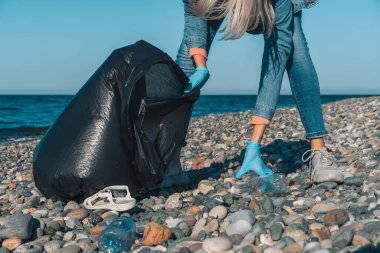 Genç bir kadın deniz kıyısındaki çakıl taşı plajında siyah bir çöp torbasında plastik topluyor. Kullanılmış kirli plastik şişeler. Karadeniz kıyısındaki çevre kirliliği