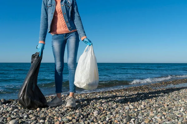 Genç bir kadın güneşli bir günde deniz kıyısındaki çakıl taşı plajında iki torba çöpü elinde tutuyor. Çevre gönüllüsü, iklim değişikliği ve okyanus kirliliğine karşı bir eylemci..