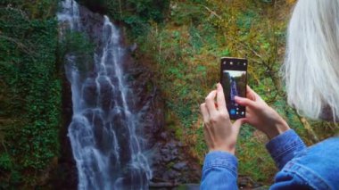 Genç bir kadın akıllı bir telefon tutuyor ve yüksek şelalenin yanında fotoğraf çekiyor.