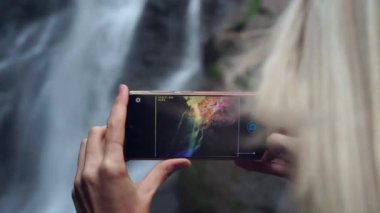 Genç bir kadın akıllı bir telefon tutuyor ve yüksek şelalenin yanında fotoğraf çekiyor.