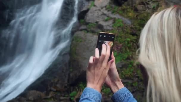 Una mujer joven sostiene un teléfono inteligente y toma fotos de pie cerca de la cascada alta — Vídeo de stock
