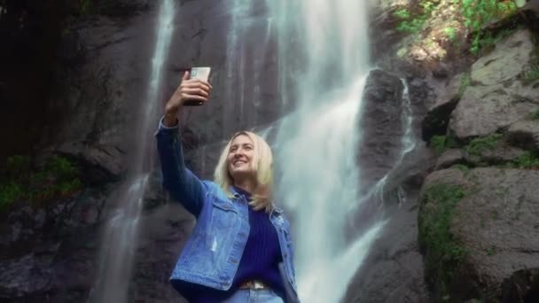 Femme tient smartphone et prend des photos d'elle-même debout près de cascade — Video