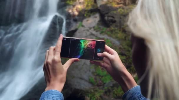 A young woman holds a smartphone and takes photos standing near high waterfall — Video Stock