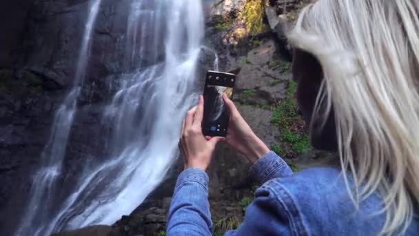 Молода жінка тримає смартфон і робить фотографії, що стоять біля водоспаду — стокове відео