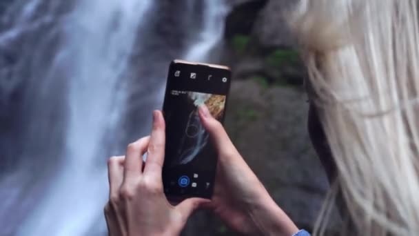 Uma jovem tem um smartphone e tira fotos em pé perto de alta cachoeira — Vídeo de Stock