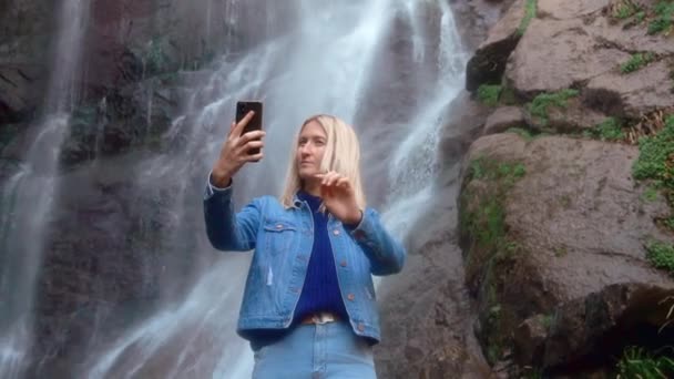 Mulher detém smartphone e tira fotos de si mesma de pé perto da cachoeira — Vídeo de Stock