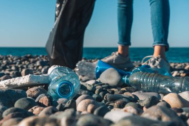 Güneşli bir günde, denizin çakıl taşı plajında siyah bir çöp torbasıyla genç bir kadının bacaklarının arka planına plastik şişelerin yakın plan çekimi. Ekoloji, gönüllü olma, çevre kirliliği kavramı.