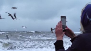 A woman takes pictures of a storm at sea and a lot of birds on her smartphone.