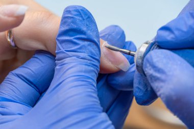 Mavi eldivenli bir manikür ustasının ellerini makrodaki bir güzellik salonundaki tırnak altlarını budamak ve sökmek için elektrikli tırnak törpüsü kullanarak yakın plan. Donanım manikürü