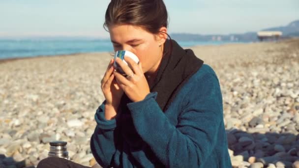 Mujer sostiene una taza de té y cuidadosamente mira profundamente en sí misma en la orilla del mar. — Vídeos de Stock