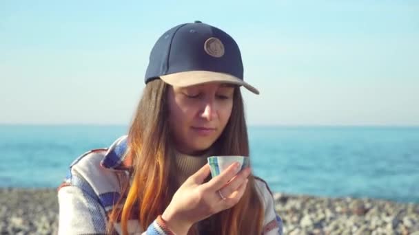 Mujer sostiene una taza de té y cuidadosamente mira profundamente en sí misma en la orilla del mar. — Vídeos de Stock