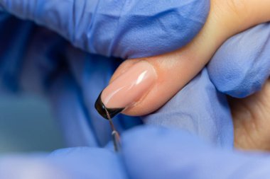 Mavi eldivenli bir manikür ustasının yakın çekimi, bir müşterinin tırnaklarına fırçayla siyah bir ceket çizer. Jöle cilası ve desenli profesyonel manikür.