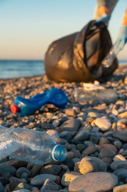 Plastik şişeleri, gün batımında denizin çakıl taşı plajında siyah çöp torbası taşıyan genç bir kadının arka planına yakın çekim. Ekoloji kavramı, gönüllülük. Dikey fotoğraf.