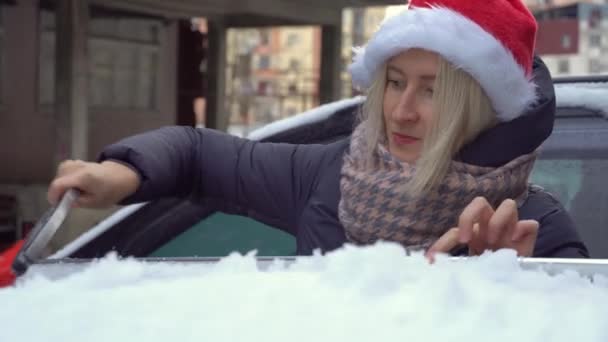 Młody piękny biały kobieta w Santa kapelusz czyści samochód z śniegu z pędzlem — Wideo stockowe