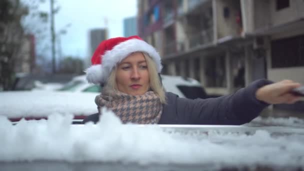 Joven hermosa mujer caucásica en Santa sombrero limpia el coche de la nieve con un cepillo — Vídeos de Stock