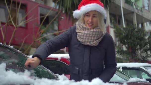 Mladá krásná běloška v santa klobouk čistí auto od sněhu s kartáčem — Stock video