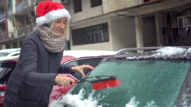 Mladá krásná běloška v santa klobouk čistí auto od sněhu s kartáčem — Stock video