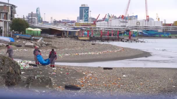 조류에 의해 해안으로 쓸려가는 플라스틱 폐기물. 두 명의 노동자가 쓰레기를 자루에 넣고 있습니다. — 비디오