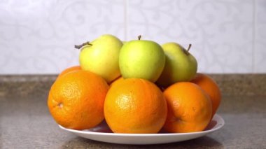 Kadının eli bir elmayı alır ve masanın üzerinde duran meyve tabağına koyar.