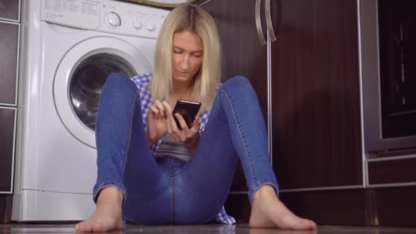 Una donna è seduta sul pavimento della cucina con il suo telefono. Bottiglie con detergente — Video Stock