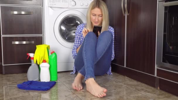 Una mujer está sentada en el piso de la cocina con su teléfono. Botellas con agente limpiador — Vídeos de Stock