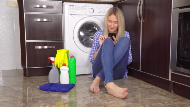 Blondine in Jeans und Hemd sitzt auf dem Küchenboden neben der Waschmaschine — Stockvideo