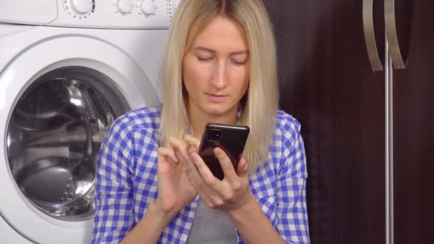 Μια γυναίκα κάθεται στο πάτωμα της κουζίνας με το τηλέφωνό της. Φιάλες με καθαριστικό — Αρχείο Βίντεο