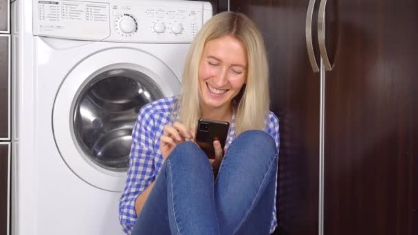 Μια γυναίκα κάθεται στο πάτωμα της κουζίνας με το τηλέφωνό της. Φιάλες με καθαριστικό — Αρχείο Βίντεο